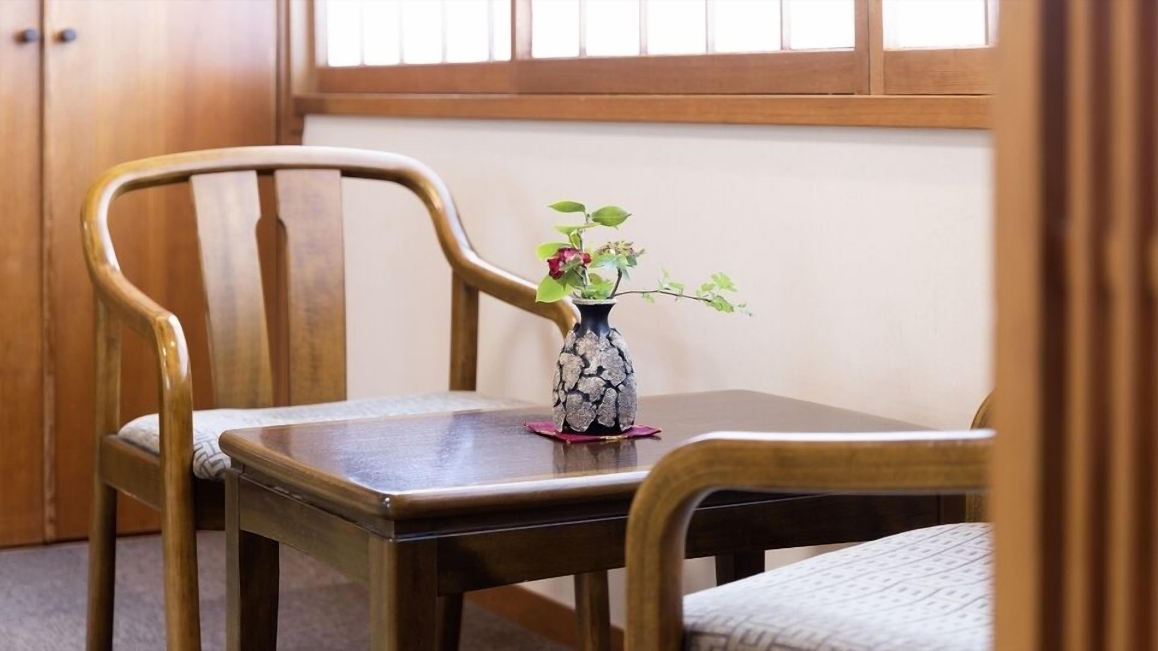 A wooden table with a vase of flowers, two wooden chairs, and a window with wooden blinds.