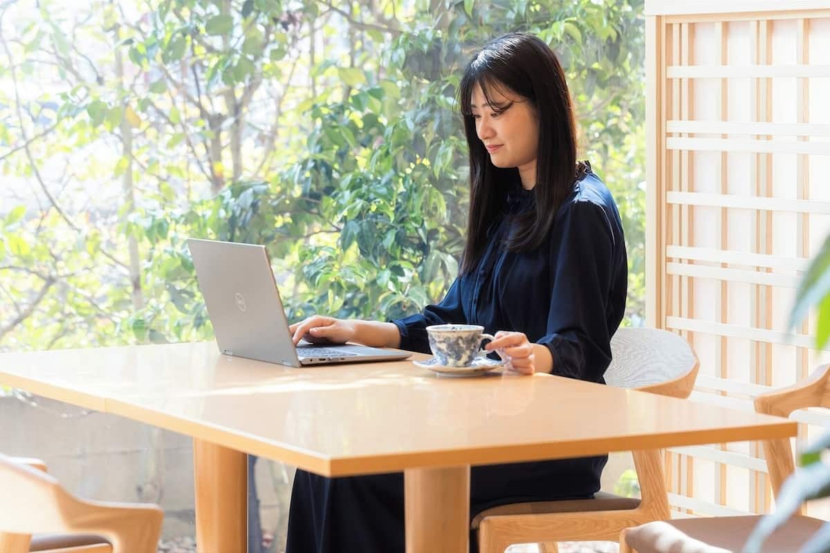 A person using a laptop at a wooden table with a cup of tea, surrounded by greenery.