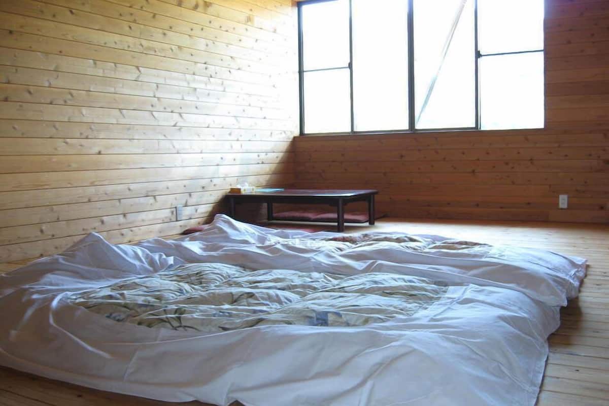 A room with a wooden floor, a bed with white bedding, a small table, and a window with vertical panes.