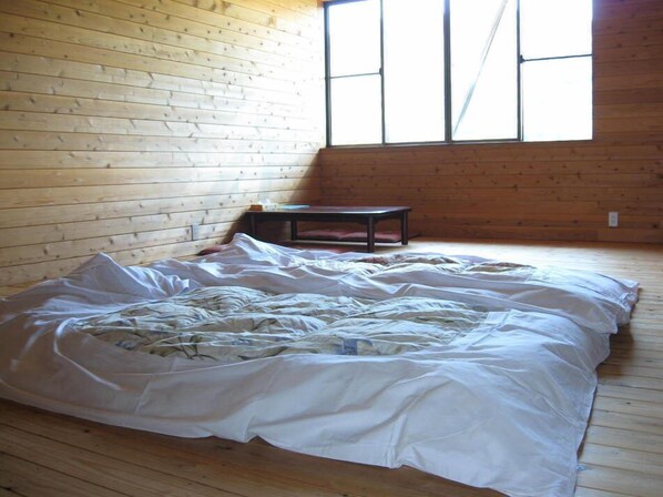 A room with a wooden floor, a bed with white bedding, a small table, and a window with vertical panes.