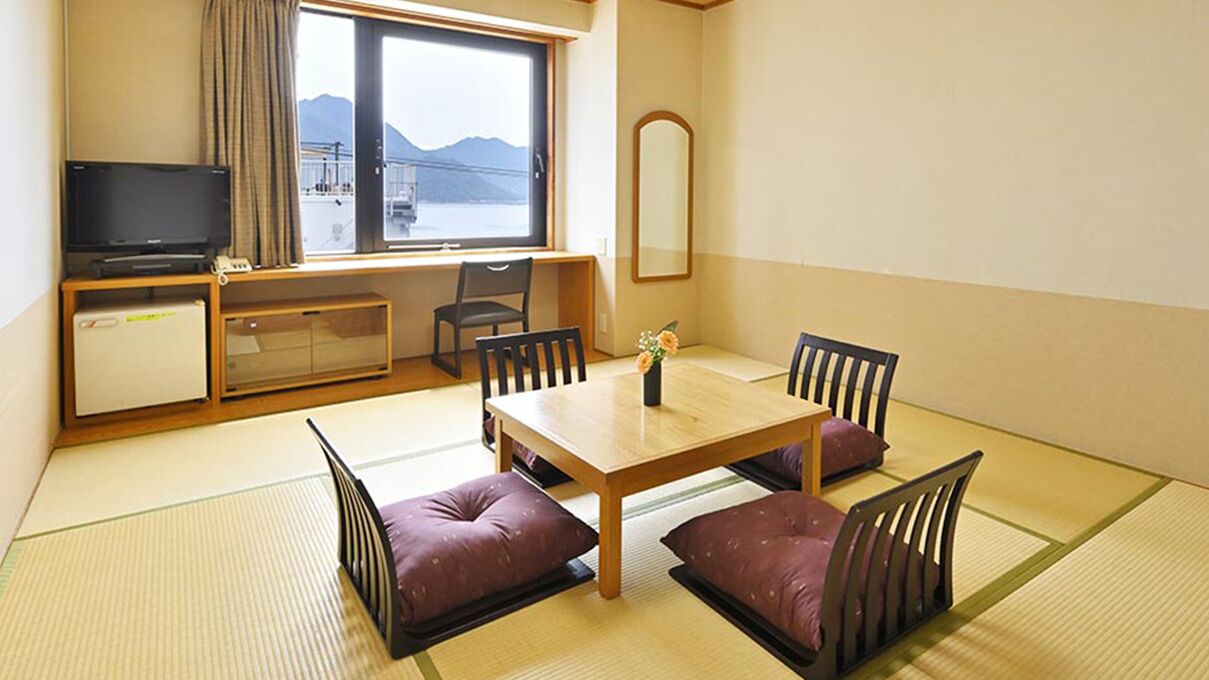 Une pièce avec un sol recouvert de tatamis, une table à manger en bois avec quatre chaises, un téléviseur posé sur un meuble, une petite armoire et une fenêtre donnant sur les montagnes.