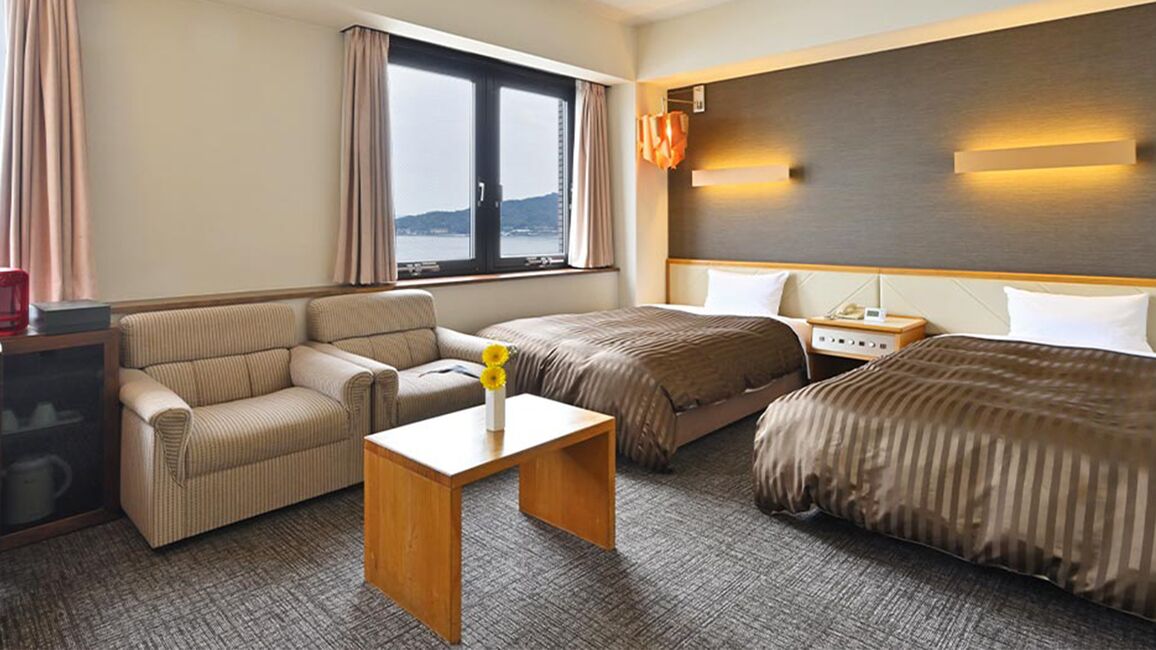 Une chambre d’hôtel avec un canapé, deux lits simples, une table de chevet, une petite table et une vue sur la mer.
