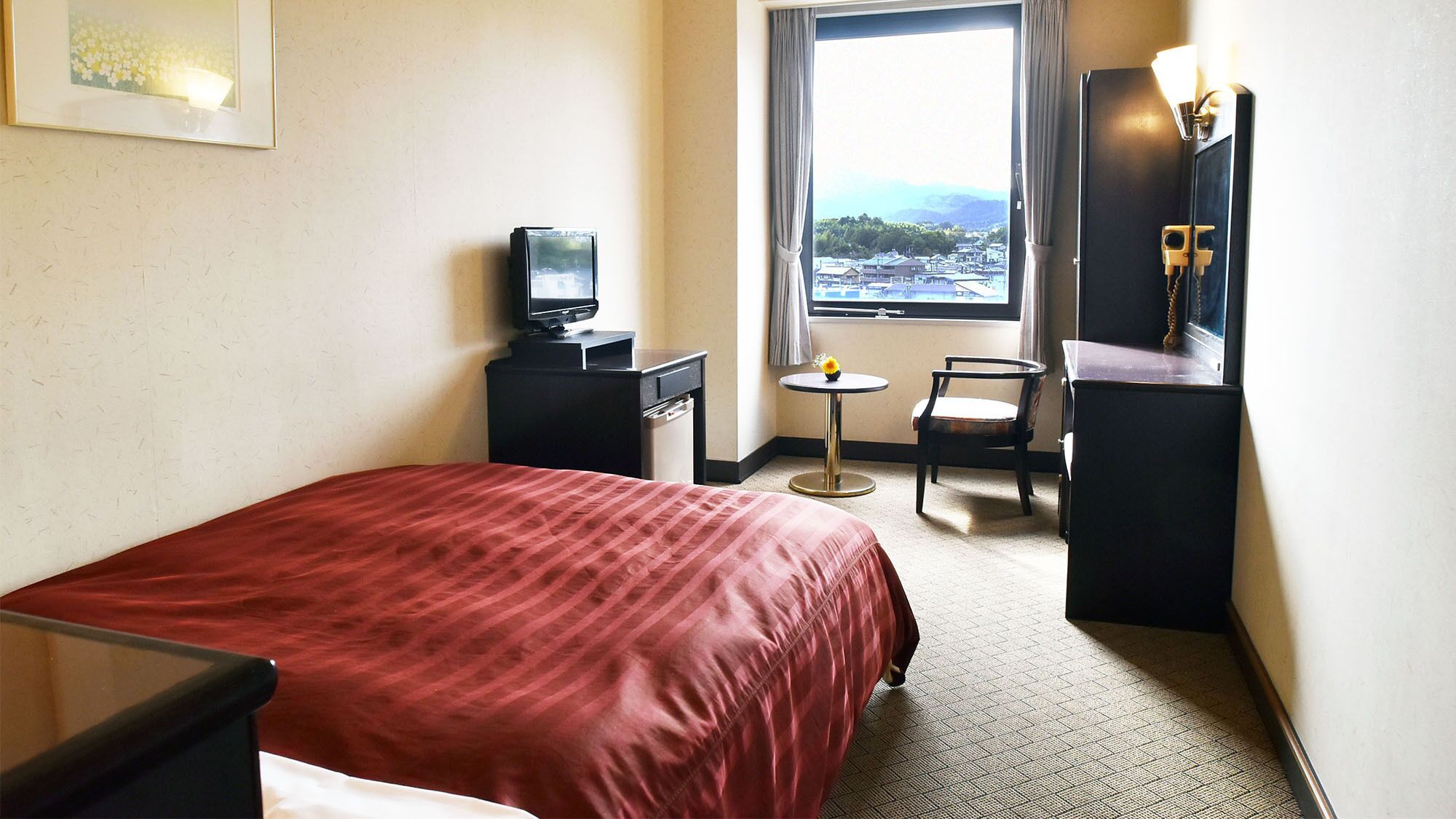 Une chambre d’hôtel avec un lit, un bureau avec un ordinateur, une chaise, une petite table avec une fleur et une fenêtre donnant sur un beau paysage.