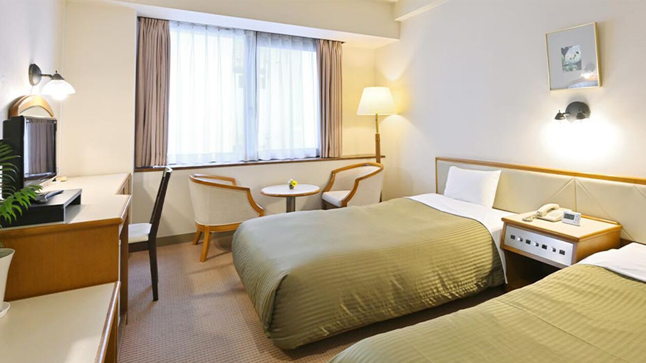 Une chambre d’hôtel comprenant un lit, une table de chevet, une lampe, une chaise, un bureau et une fenêtre avec des rideaux.