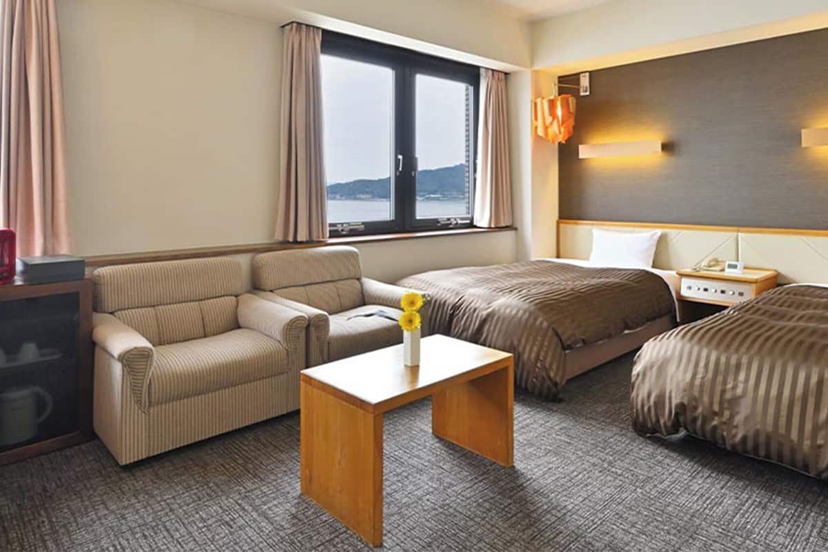 Hotellihuone, jossa on sohva, kaksi erillistä sänkyä, pieni pöytä maljakolla, televisio ja ikkuna, jossa on verhot.