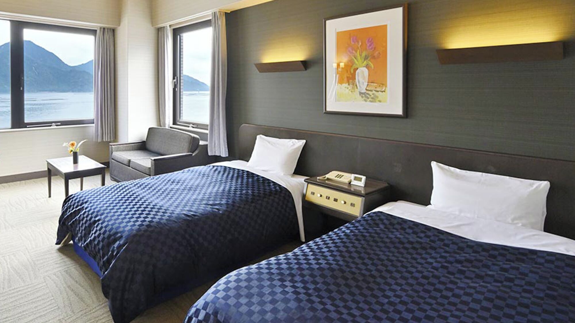 Hotellihuone, jossa on kaksi sänkyä, työpöytä, tuoli, yöpöytä, taulu ja ikkuna, josta avautuu näkymä merelle ja vuorille.