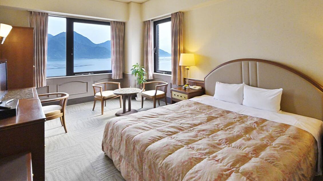 Une chambre d’hôtel avec un grand lit, un bureau, une chaise, une petite table et une table de chevet. Il y a deux fenêtres avec des rideaux, et une plante est posée sur la table de chevet.