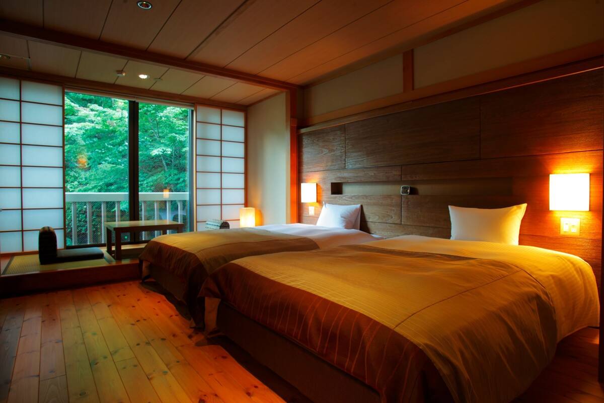 A hotel room with two beds, a wooden floor, a large window with a view of trees, and a wooden headboard.