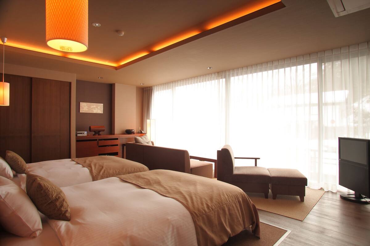 A hotel room with two beds, a desk, a television, and large windows.