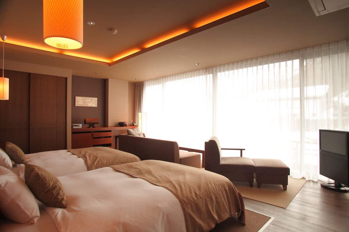 A hotel room with two beds, a desk, a television, and large windows.