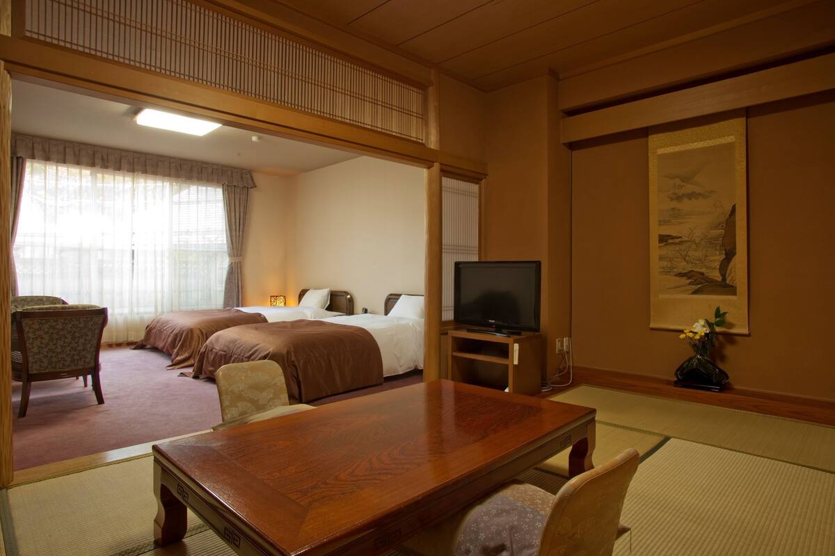A hotel room with a bed, a television, a dining table, and a traditional Japanese artwork on the wall.