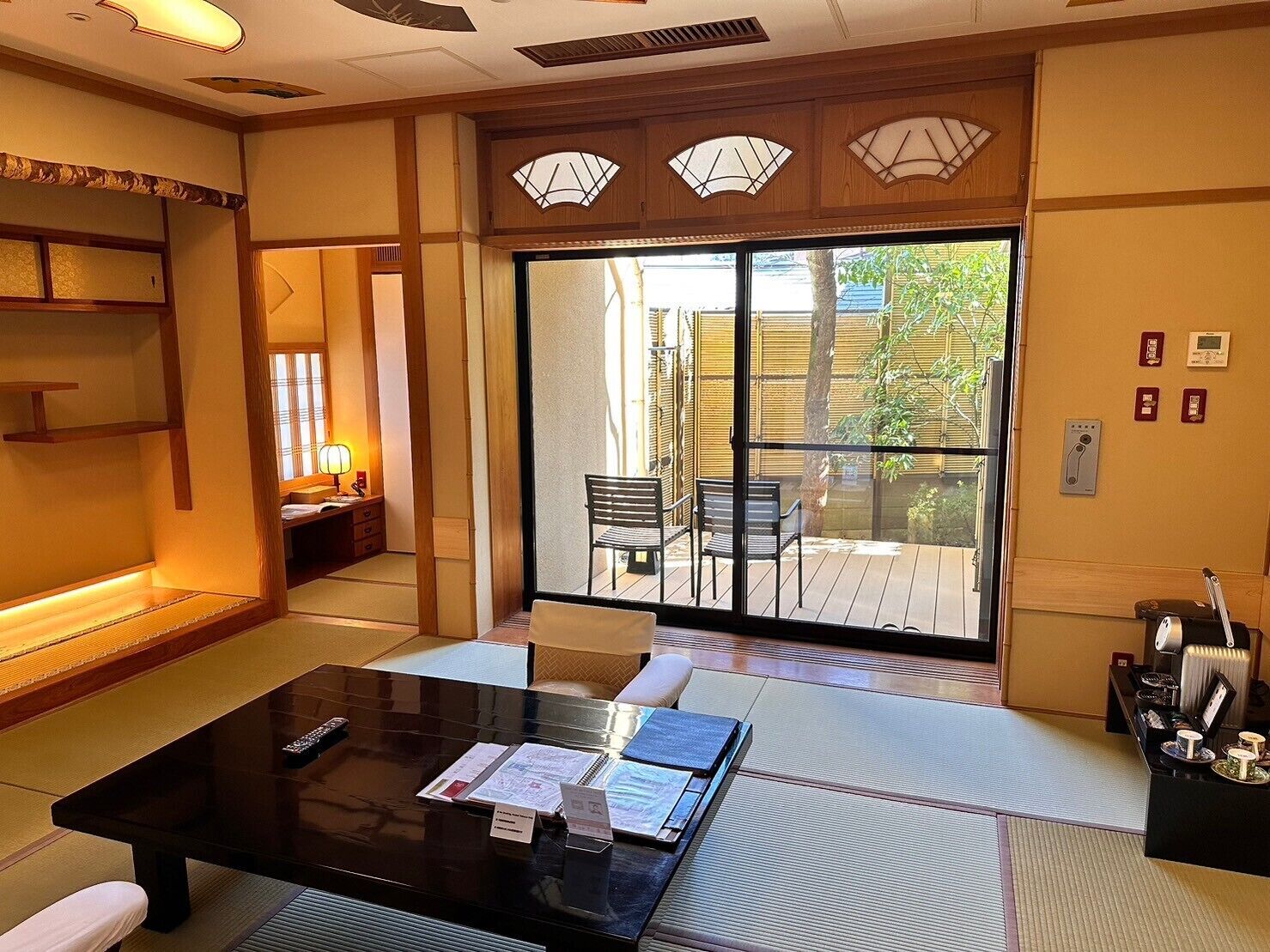 A traditional Japanese-style room with tatami flooring, a low table with papers, a sliding door leading to an outdoor area with chairs, and a small desk with a telephone and a radio.