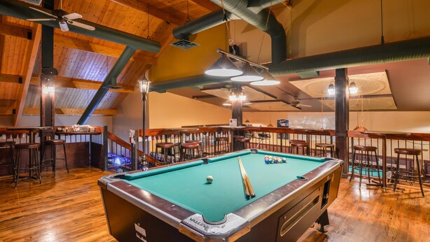 Une table de billard se trouve dans une pièce au plancher en bois et au plafond haut. On aperçoit des tabourets de bar et un comptoir de bar à l’arrière-plan.