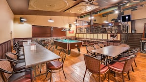 Une salle de billard équipée de tables, de chaises, d’un billard américain, de ventilateurs de plafond et d’un coin bar avec des tabourets.