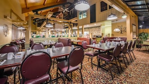 Un restaurant avec de hauts plafonds, des poutres en bois et un tapis à motifs. Les tables sont dressées avec des nappes blanches et des chaises rouges.