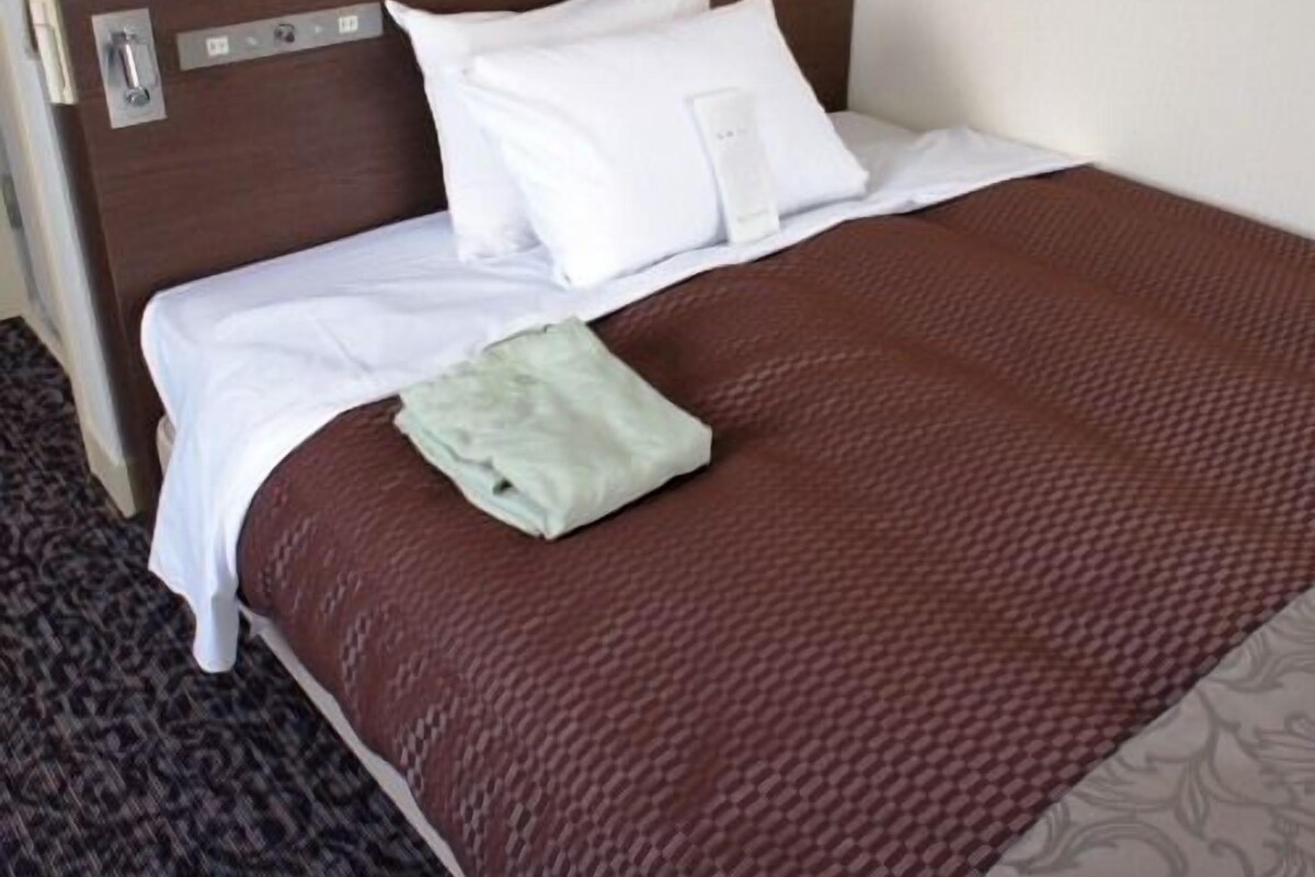 A neatly made bed with white linens and a brown bedspread, a bedside telephone, and a small bedside table with a tissue box.