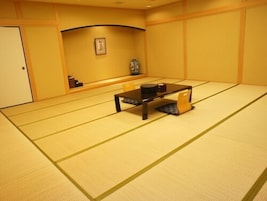 Una habitación con esterillas de tatami, una mesa baja y cojines, un cuadro en la pared y un pequeño estante con objetos.