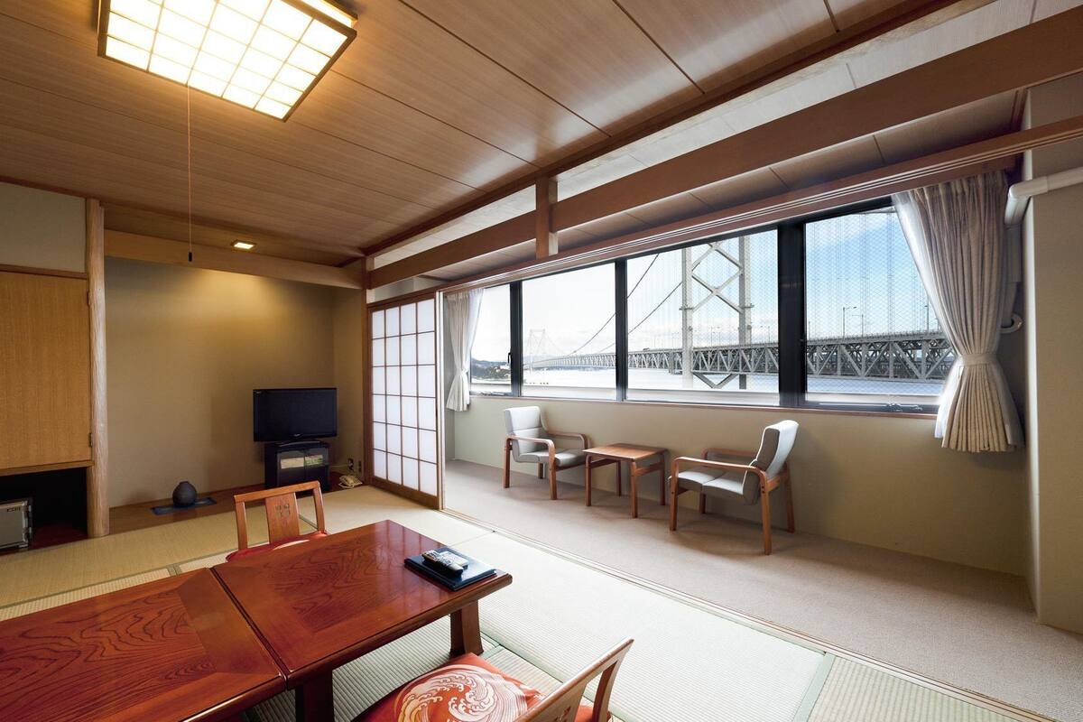 A traditional Japanese room with tatami flooring, a low table, chairs, a television, and a view of a bridge.