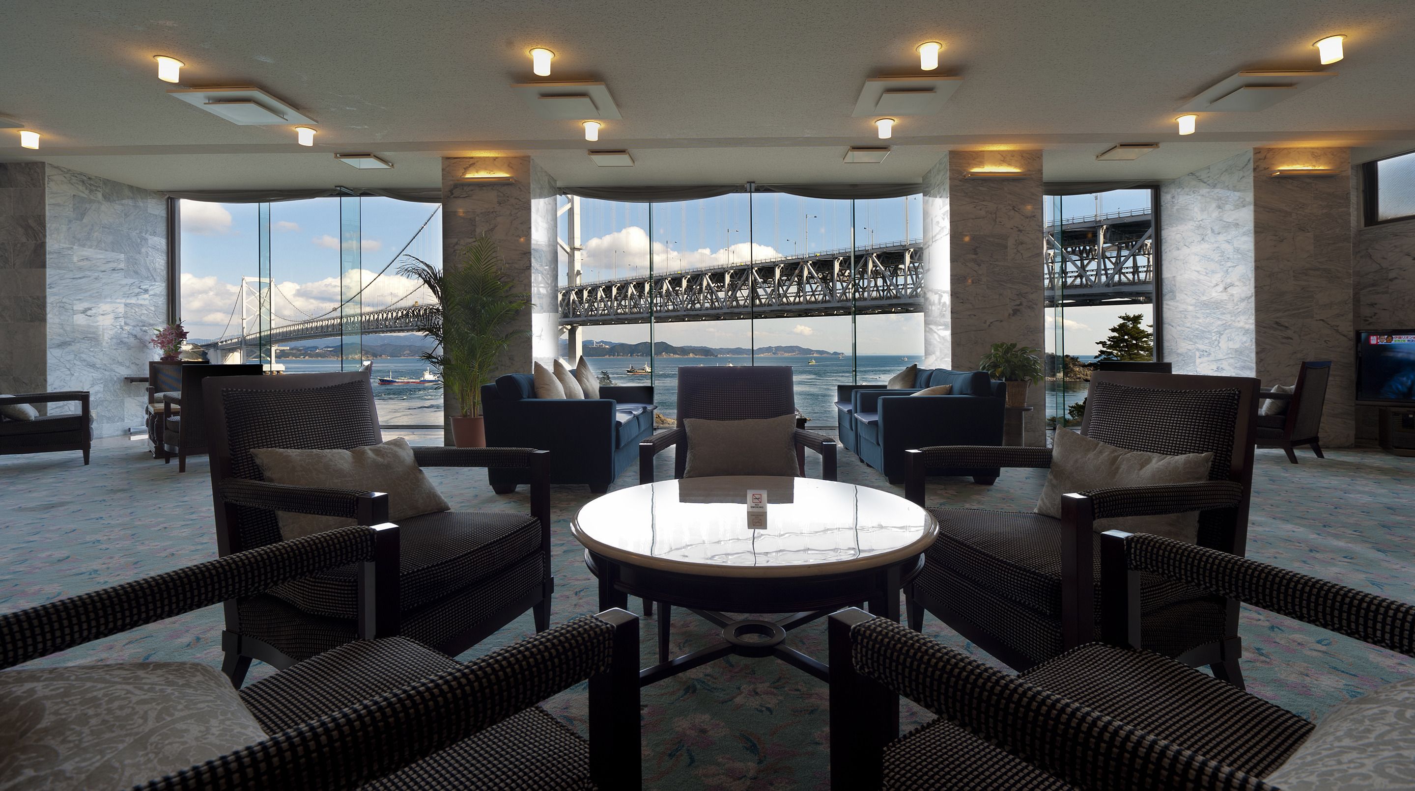 A spacious lounge area with a large round table, comfortable seating, and a view of a bridge and water.