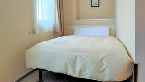 Una cama pequeña y bien hecha en una habitación con un único cuadro en la pared y una ventana con cortinas traslúcidas.