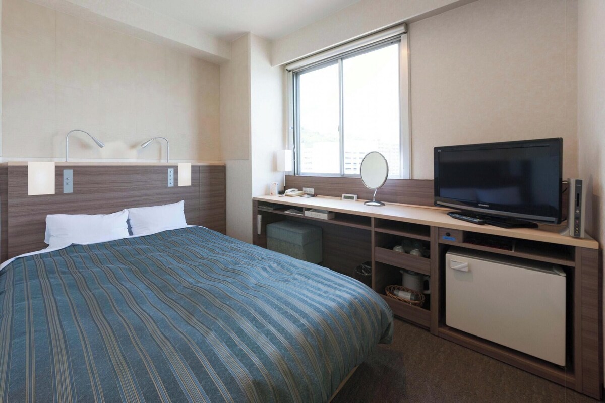Une chambre d’hôtel équipée d’un lit, d’une télévision, d’un bureau avec un ordinateur, d’un miroir et d’une fenêtre.
