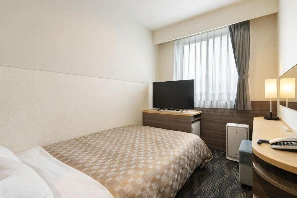 Une chambre d’hôtel avec un lit, une télévision, un bureau avec une lampe et une fenêtre avec des rideaux.
