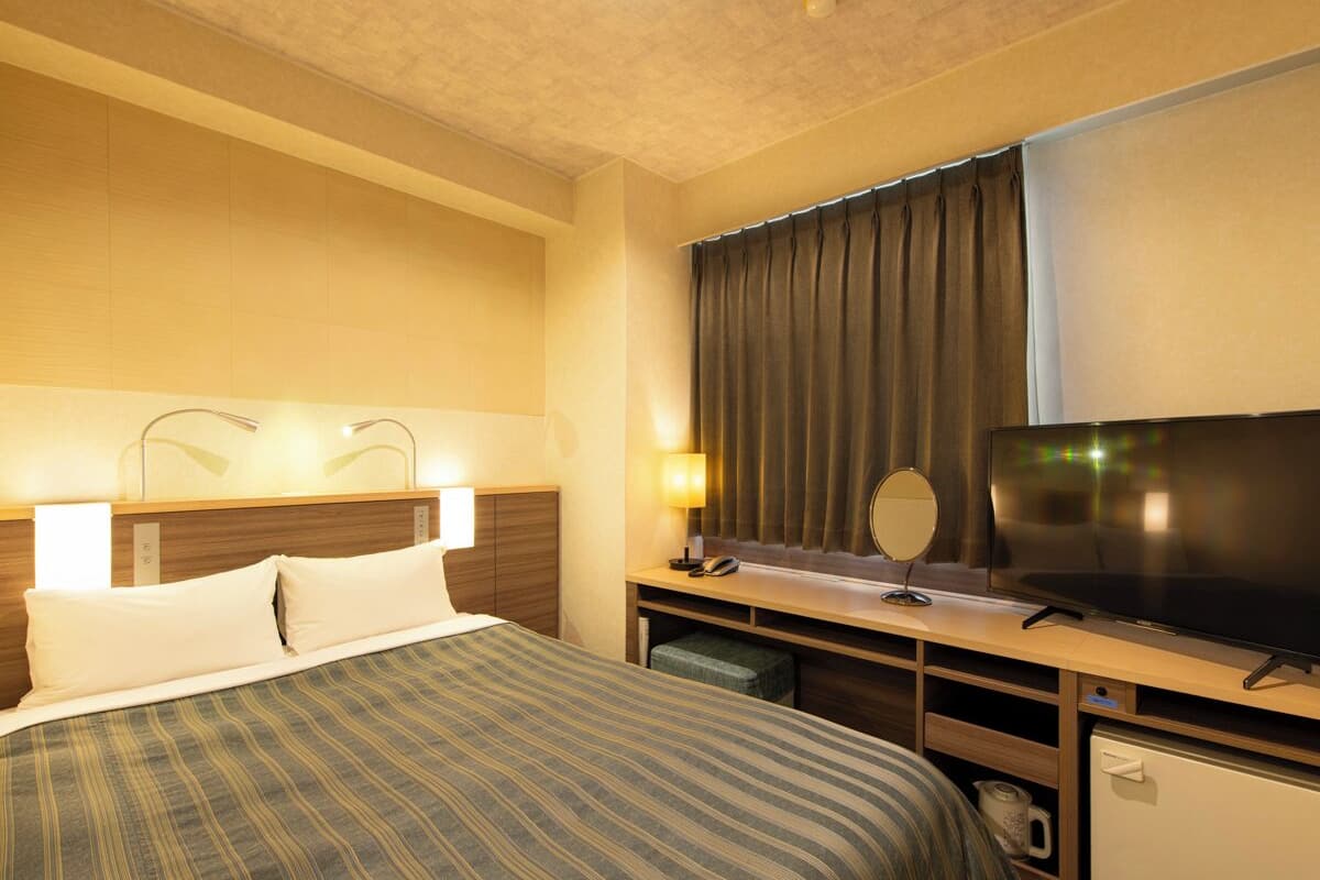 A hotel room with a bed, a desk with a television, a mirror, and a small refrigerator.