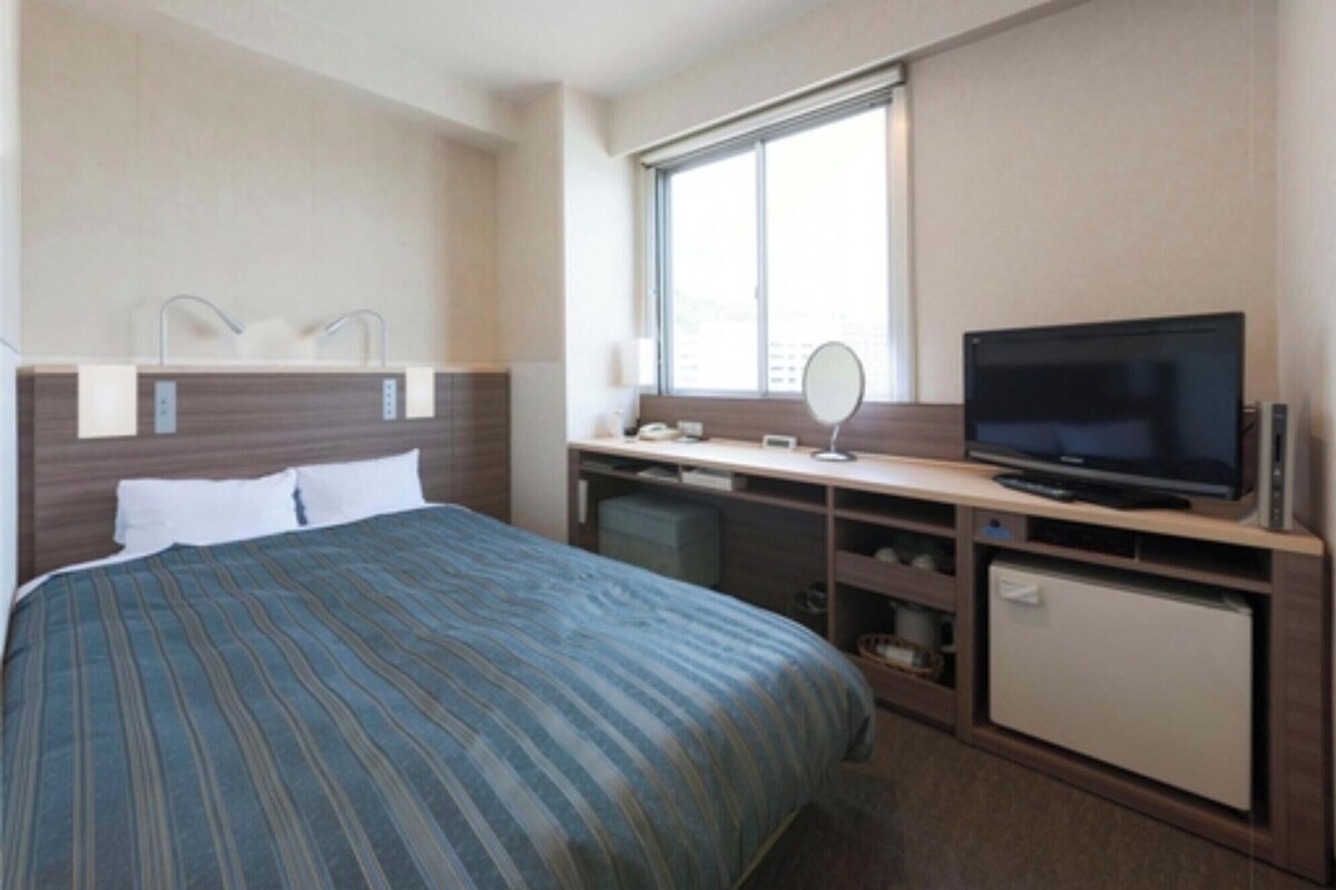 Une chambre d’hôtel avec un lit, un bureau avec une télévision, un miroir et une fenêtre.