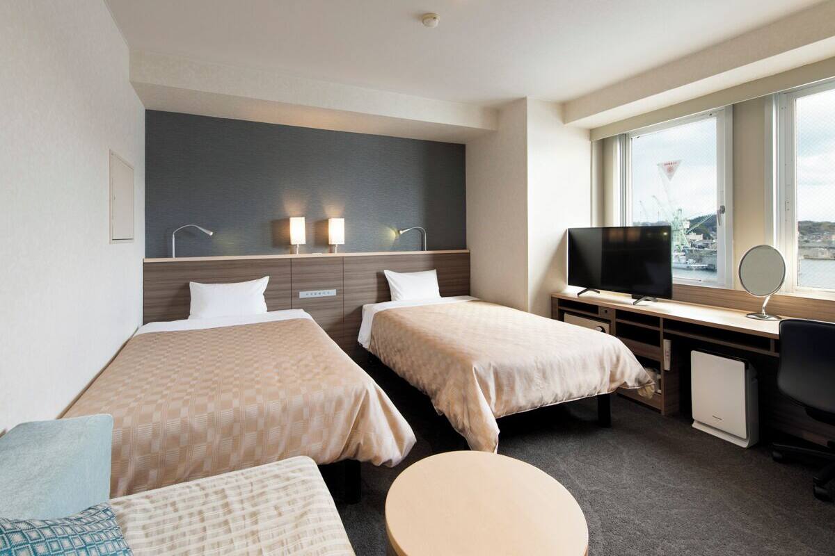 A hotel room with two beds, a desk with a computer, a TV, and a window with a view of the sea.