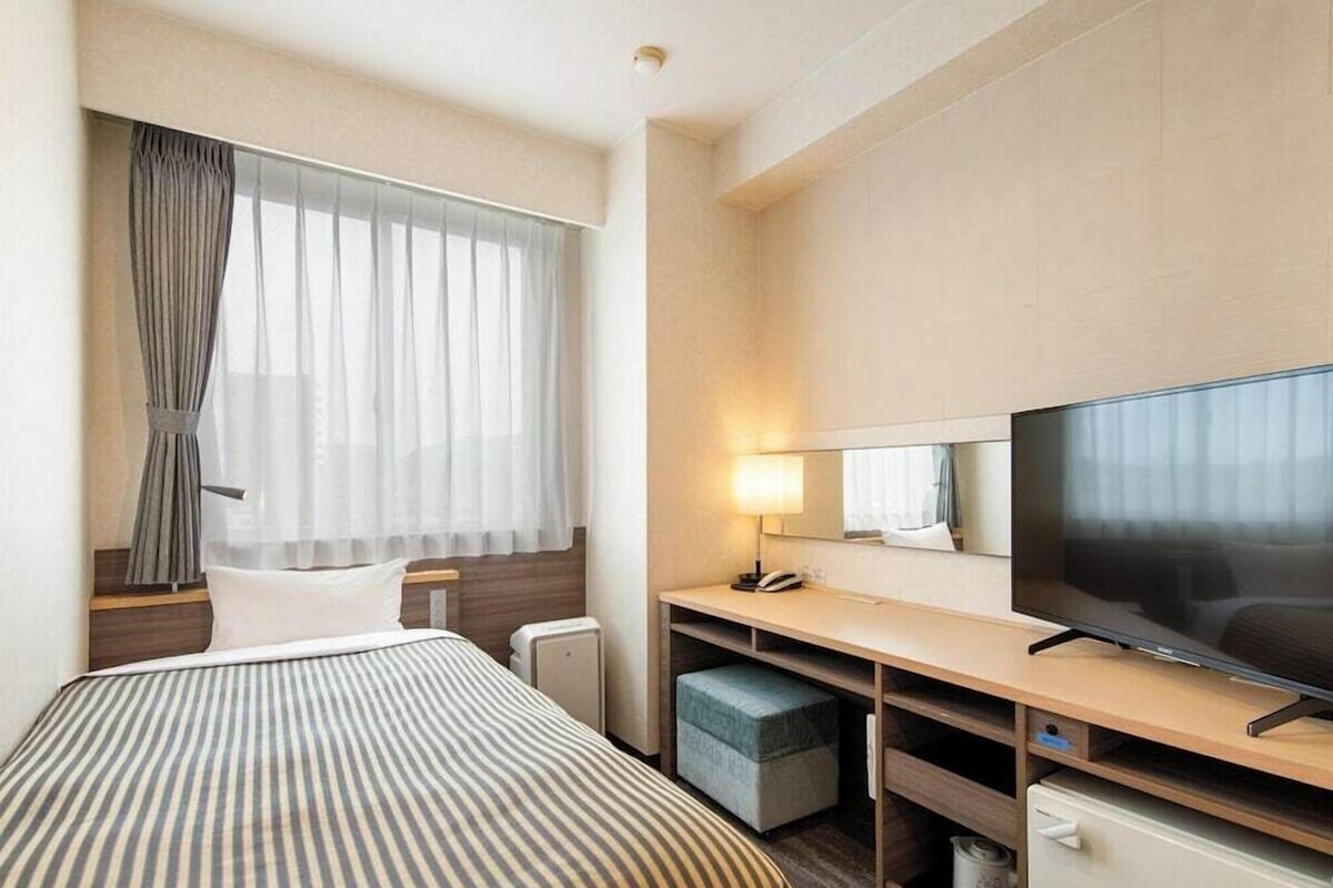 Une chambre d’hôtel avec un lit, un bureau doté d’une télévision, une lampe, un miroir et une petite poubelle.