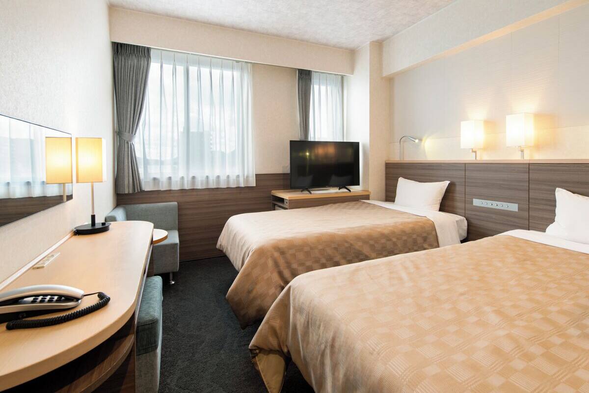 A hotel room with two beds, a desk with a telephone, a television, and a window with curtains.