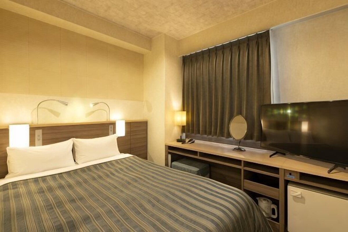 Une chambre d’hôtel comprenant un lit, des lampes de chevet, un bureau avec un miroir, une télévision et un petit réfrigérateur.
