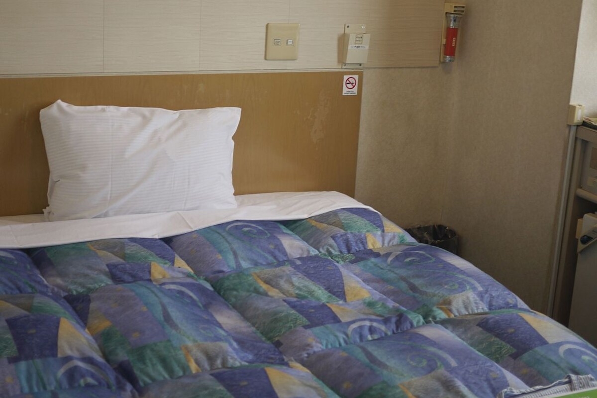 Une chambre d’hôtel comprenant un lit, une tête de lit, un interrupteur fixé au mur, un détecteur d’incendie et une poubelle.