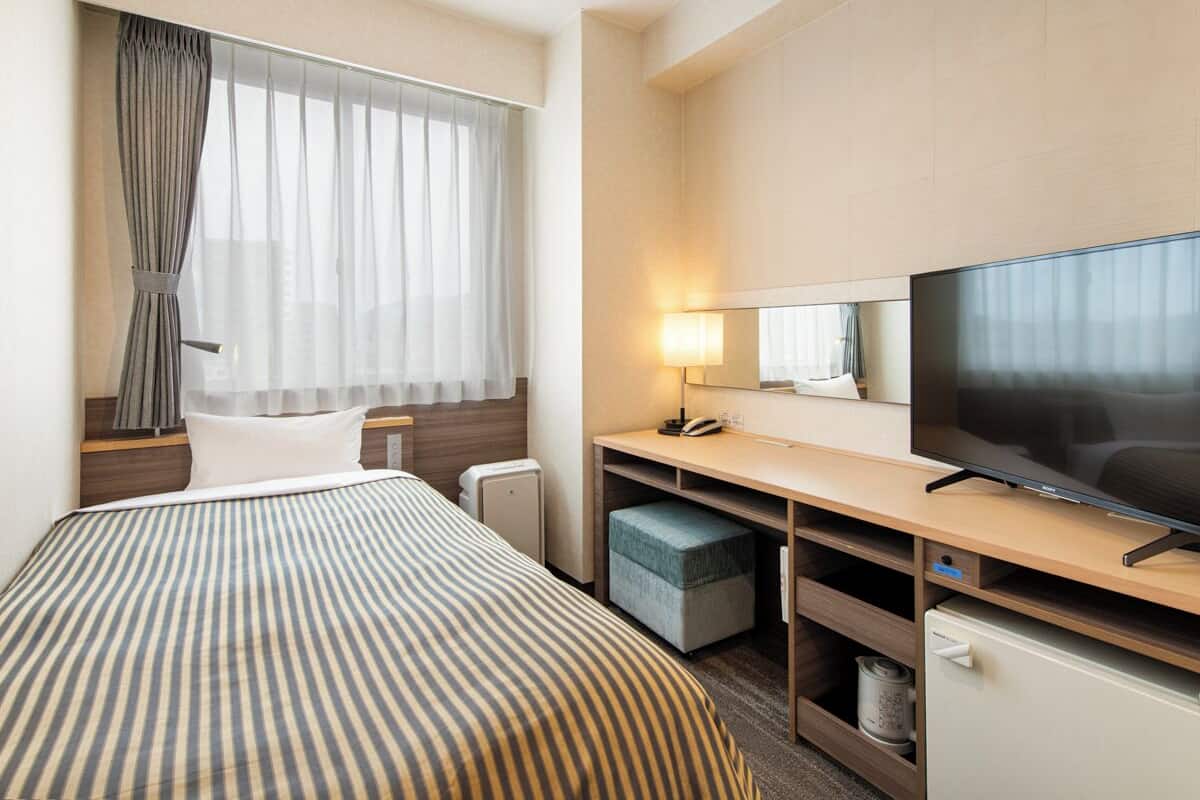 A hotel room with a bed, a desk with a TV, a lamp, and a small storage unit.