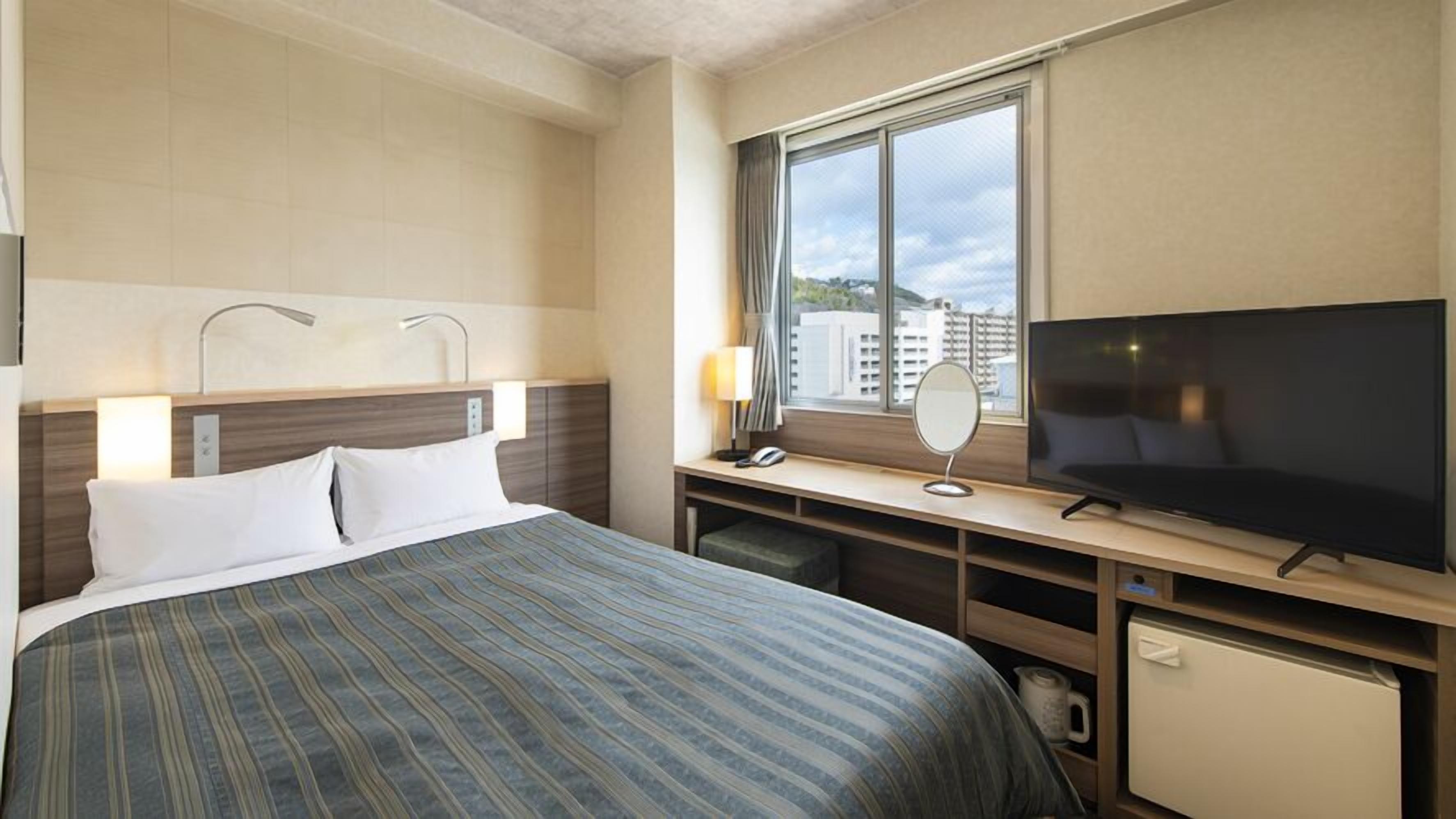 Habitación de hotel con cama, televisión, escritorio con lámpara y ventana con vista a edificios.