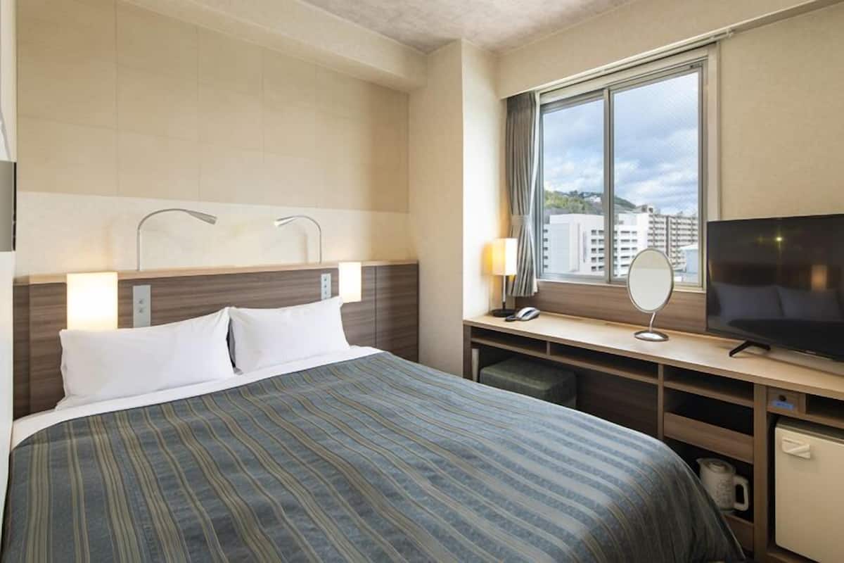 A hotel room with a bed, a TV, a desk with a lamp, and a window with a view of buildings.