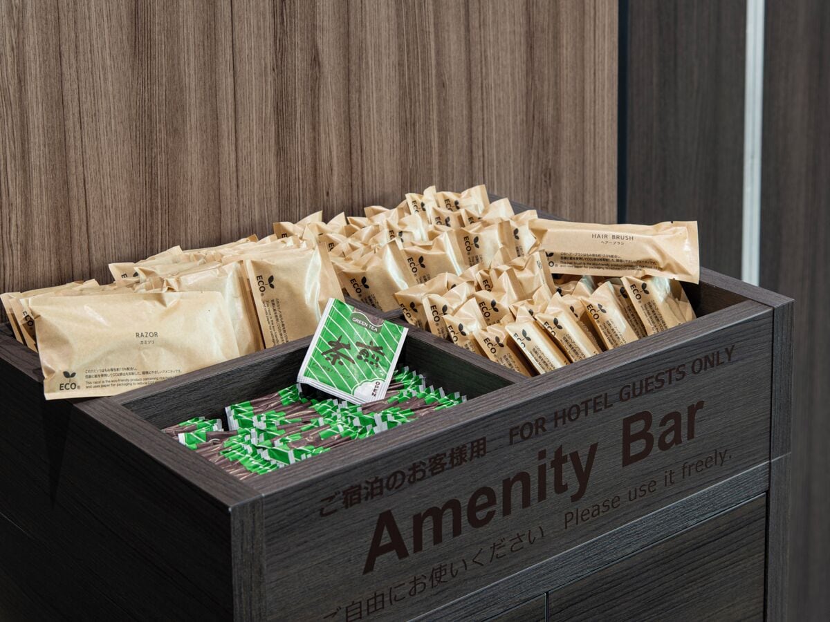 En trälåda märkt med texten ”Amenity Bar”, som innehåller olika förpackade varor, bland annat ett grönt paket och flera bruna papperspåsar.