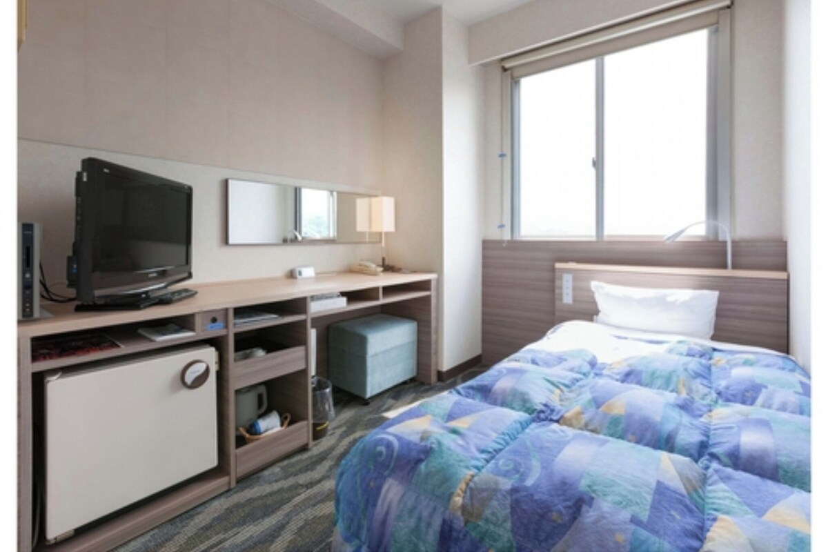 Une chambre d’hôtel comprenant un lit, un bureau avec une télévision, un petit réfrigérateur, une table de chevet et une fenêtre.