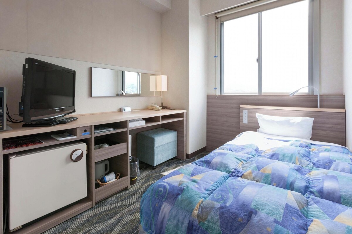 Une chambre d’hôtel comprenant un lit, un bureau avec une télévision, un miroir, une fenêtre et un petit espace de rangement.