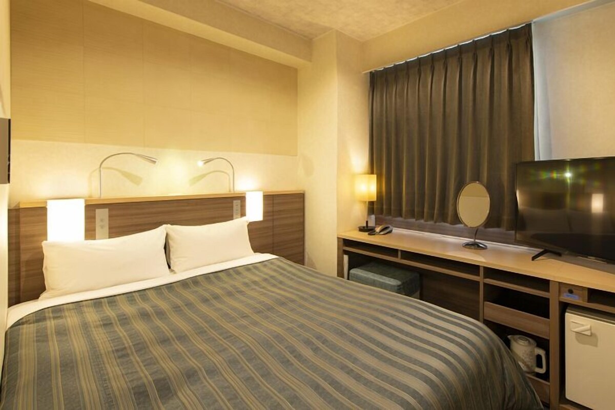 Une chambre d’hôtel équipée d’un lit, d’une télévision, d’un bureau avec une lampe, d’un miroir et d’un petit réfrigérateur.