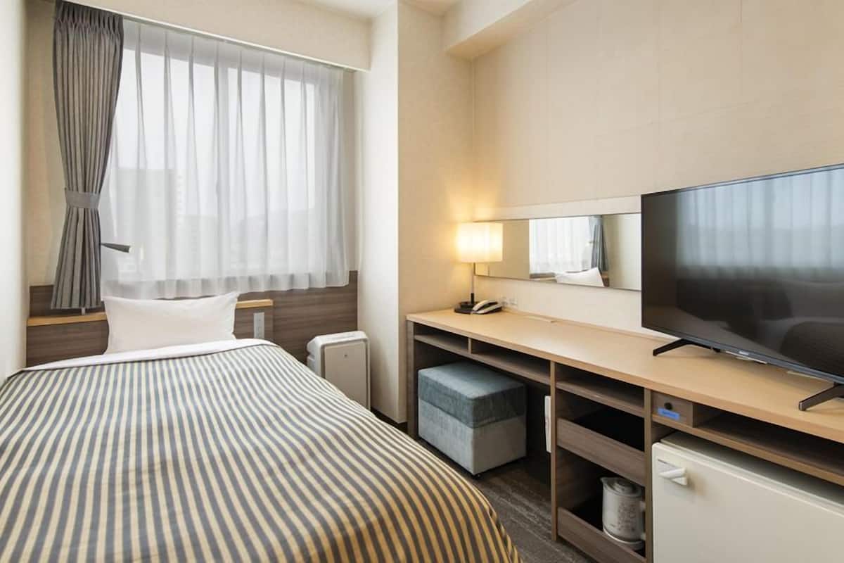 A hotel room with a bed, a desk with a TV, a mirror, and a window with curtains.