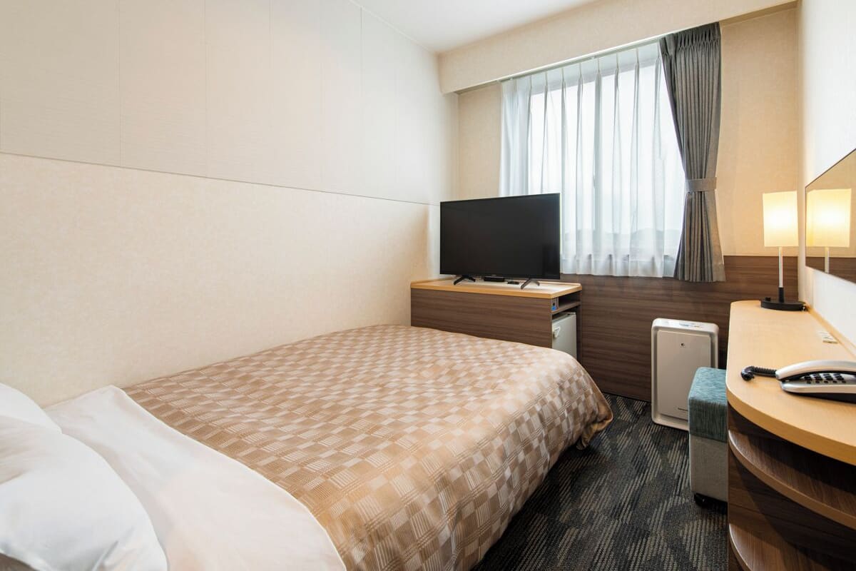 A hotel room with a bed, a desk with a flat-screen TV, a bedside table with a lamp, and a small seating area.