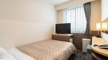 A hotel room with a bed, a desk with a flat-screen TV, a bedside table with a lamp, and a small seating area.
