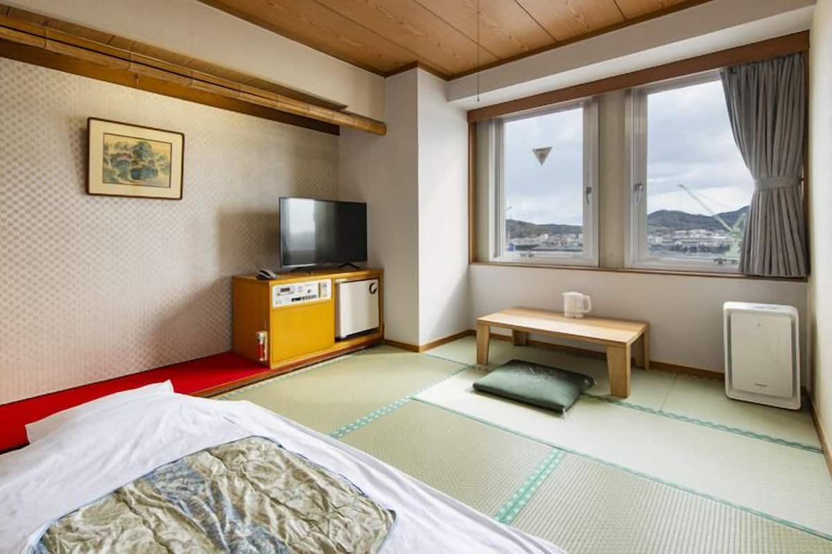 A traditional Japanese room with tatami flooring, a futon bed, a television, a small cabinet, a window with a view, and a heater.