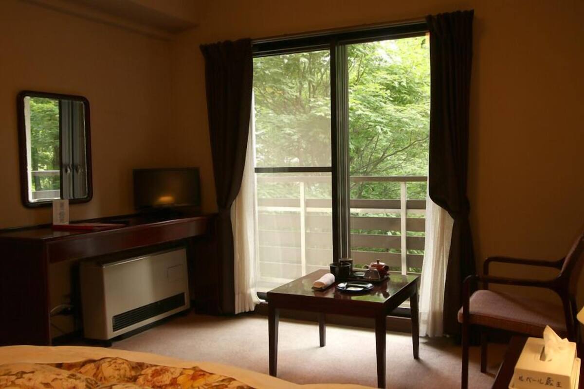 A hotel room with a bed, a desk with a television, a table with a lamp, and a view of greenery through the window.