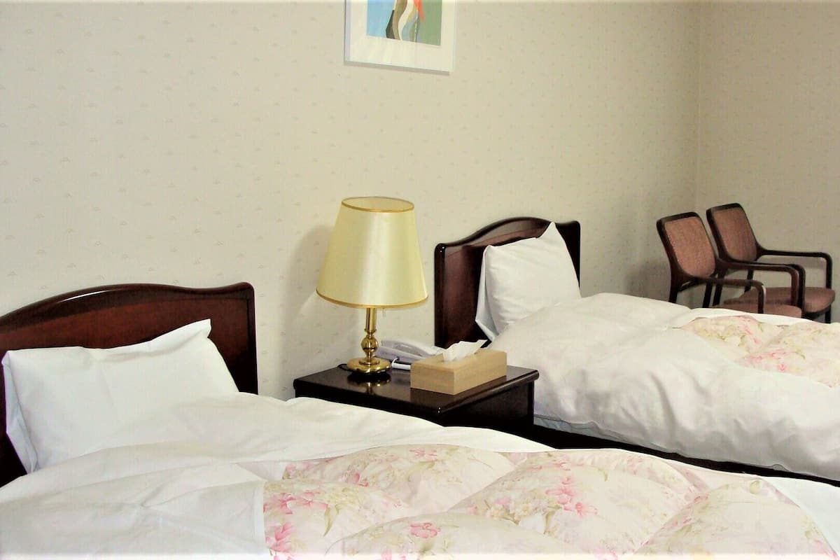 A hotel room with two beds, a bedside table with a lamp, a chair, and a small table.
