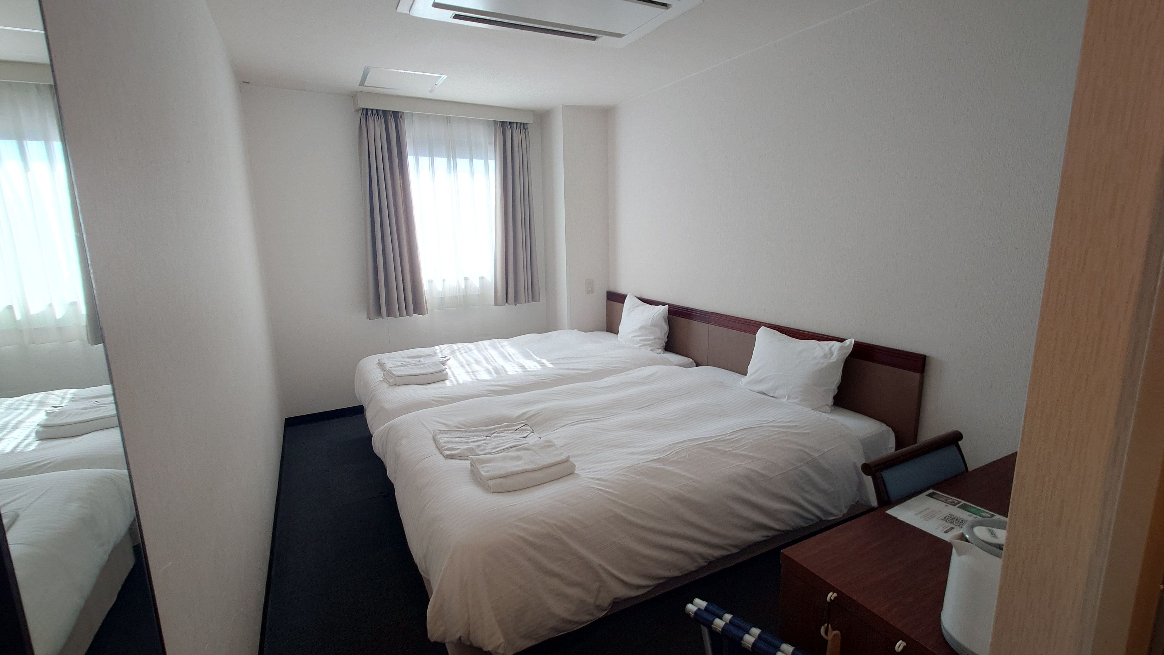 A hotel room with a double bed, a bedside table, a window with curtains, and a small desk with a chair.