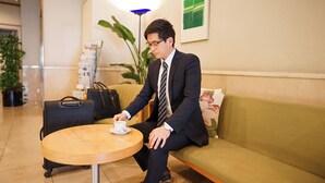 A man sitting on a green sofa in a lobby with a wooden table in front of him. There is a black suitcase and a black bag on the floor beside the sofa. - Hotel Dorf Shizuoka (Shizuoka)