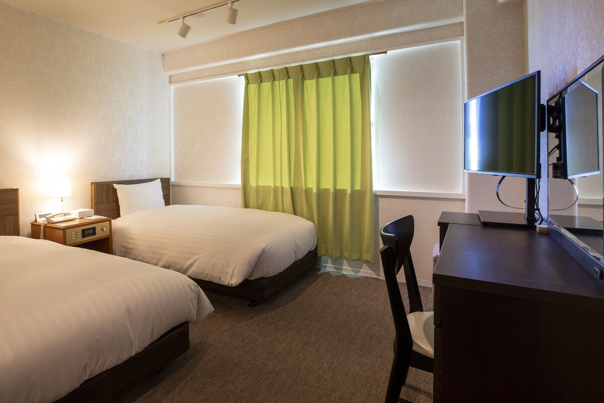A hotel room with two beds, a desk with a chair, a television, and a window with curtains.