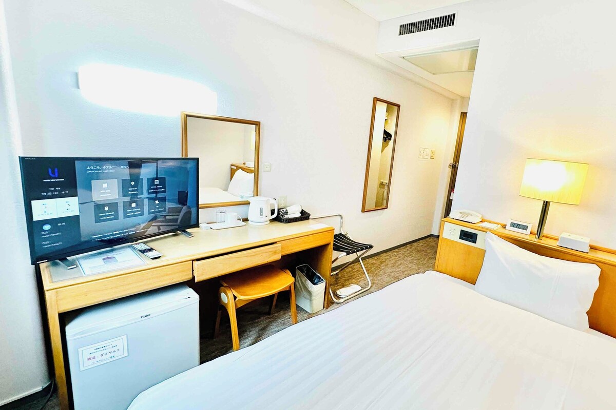 A hotel room with a bed, desk, chair, TV, and a bedside table with a lamp.